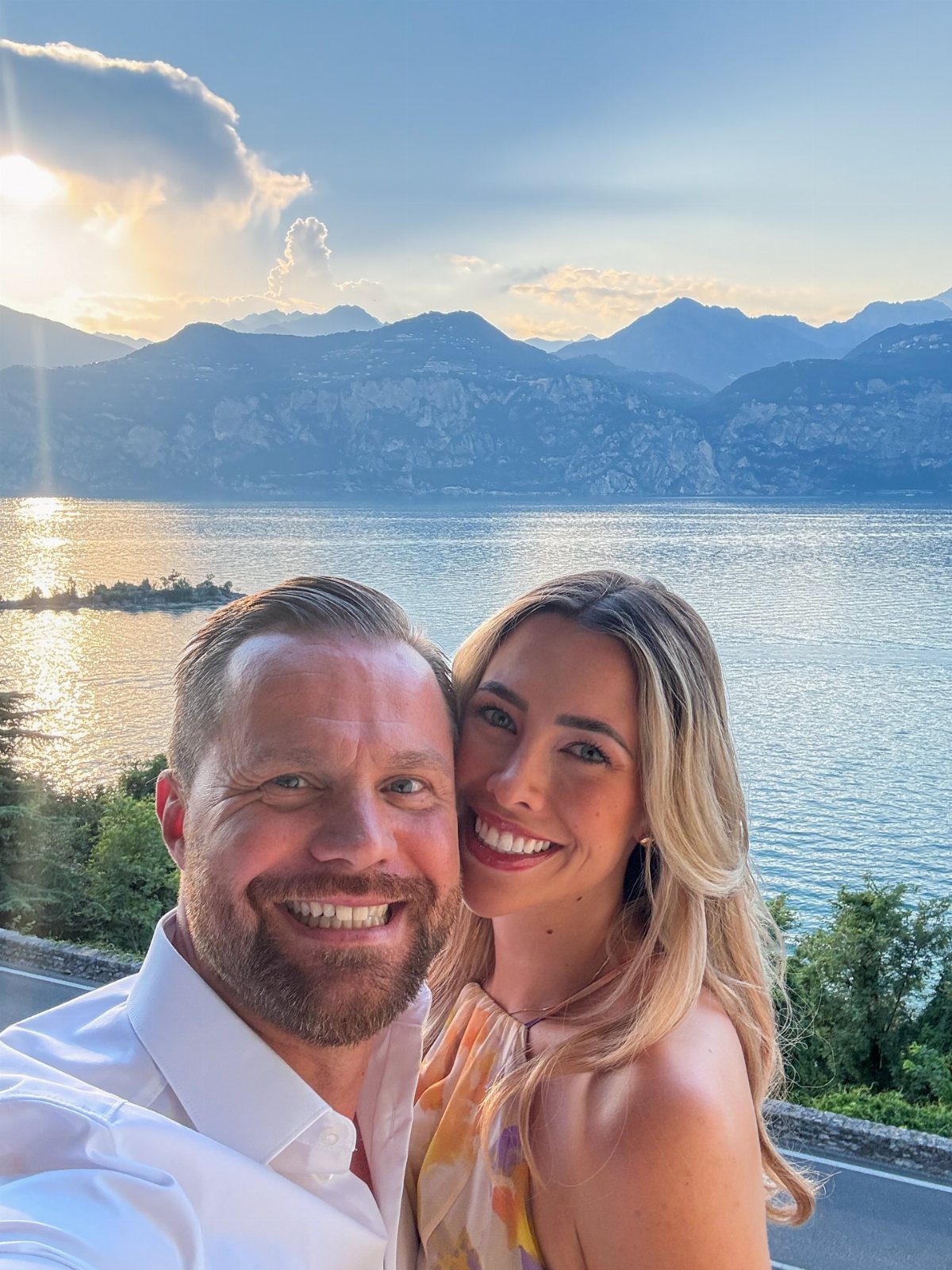 Smiling couple posing for a selfie with a scenic lake and mountains behind them.