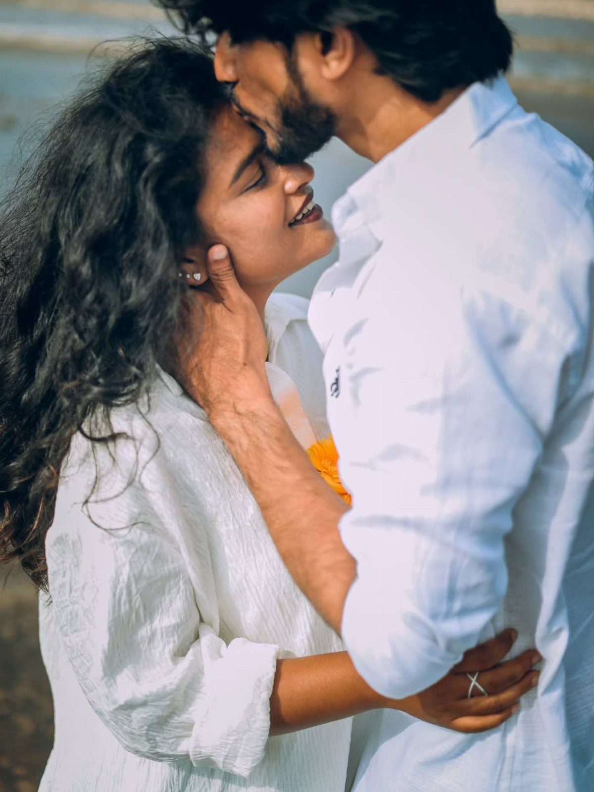 Man kissing woman's forehead while she smiles, both wearing white clothing outdoors.