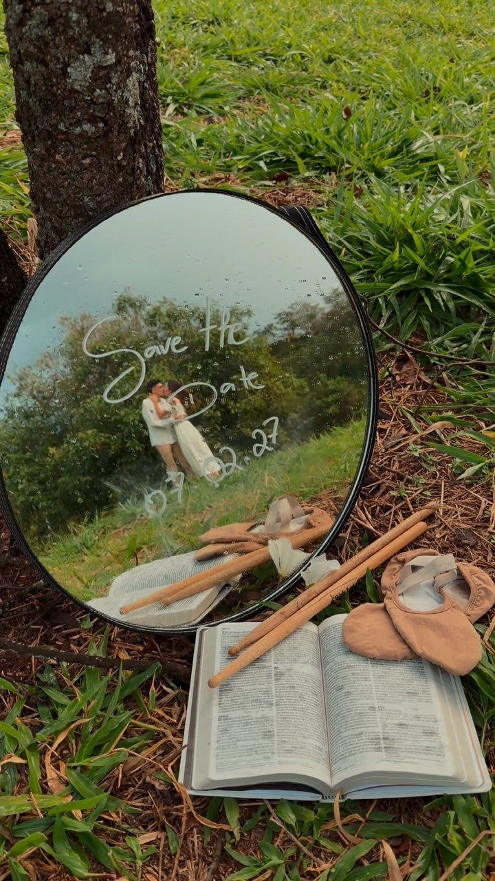Round mirror reflecting a couple embracing, placed on grass with an open book.