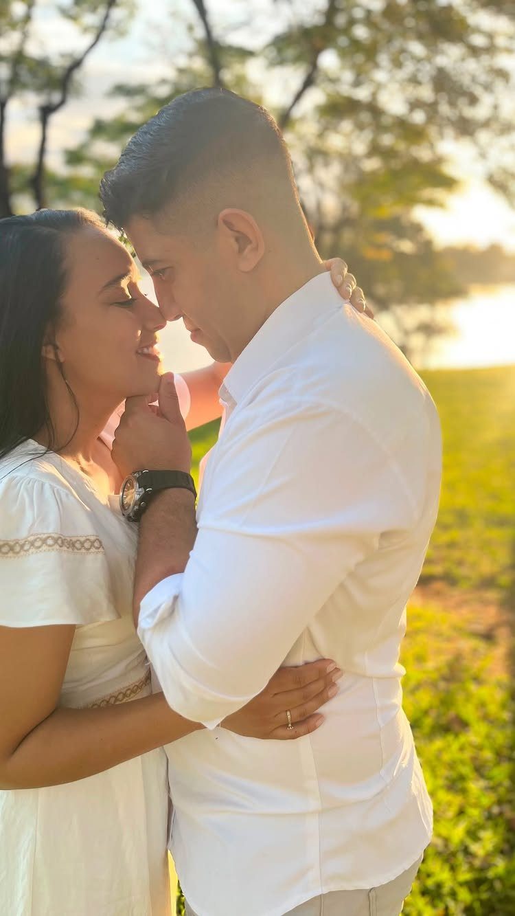 Couple in white clothing smiling and embracing in a sunny outdoor park setting.