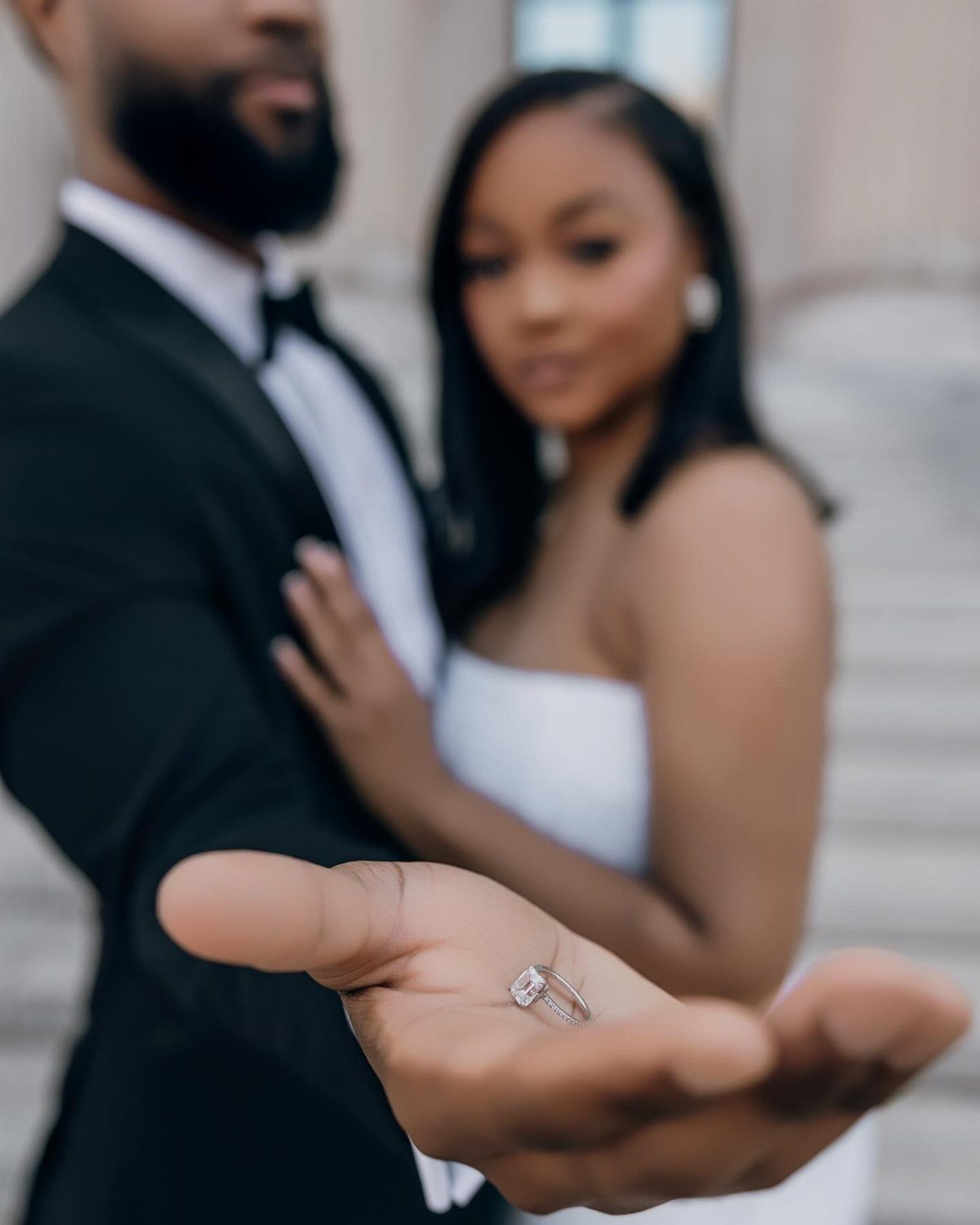 Diamond engagement ring held in a hand with a blurred couple behind.