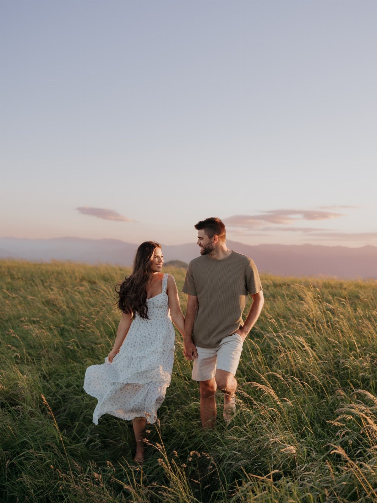 Couple holding hands and walking through a grassy field during a golden sunset.