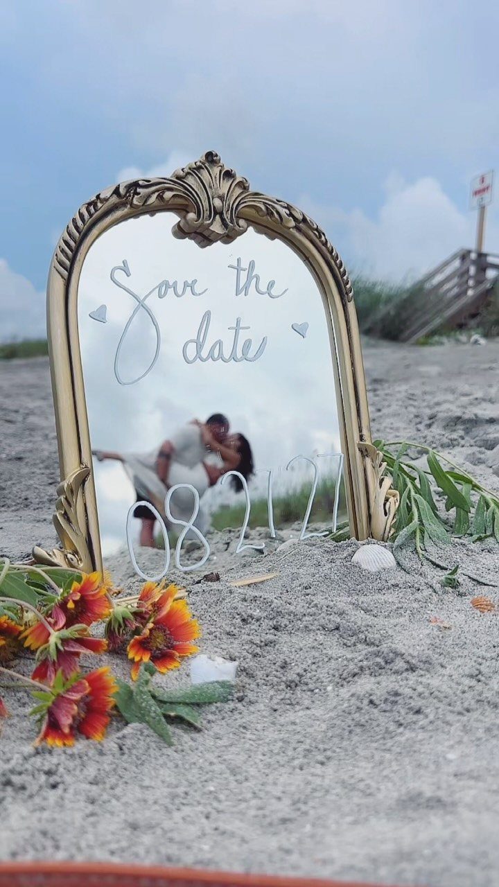 Ornate gold mirror on sand reflecting a couple with save the date calligraphy