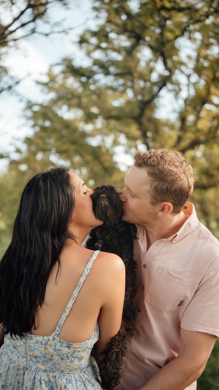 Couple kissing a small black fluffy dog outdoors with green trees behind them