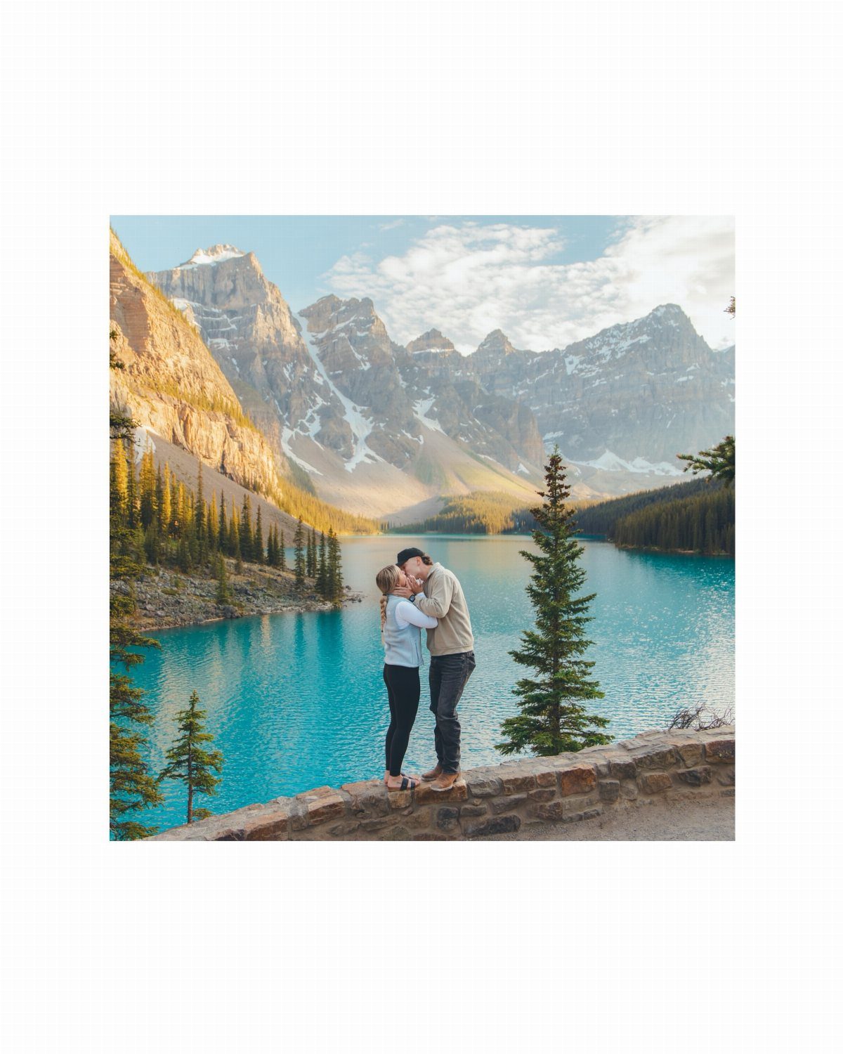 Couple kissing on a stone ledge overlooking a turquoise lake and snowy mountains.
