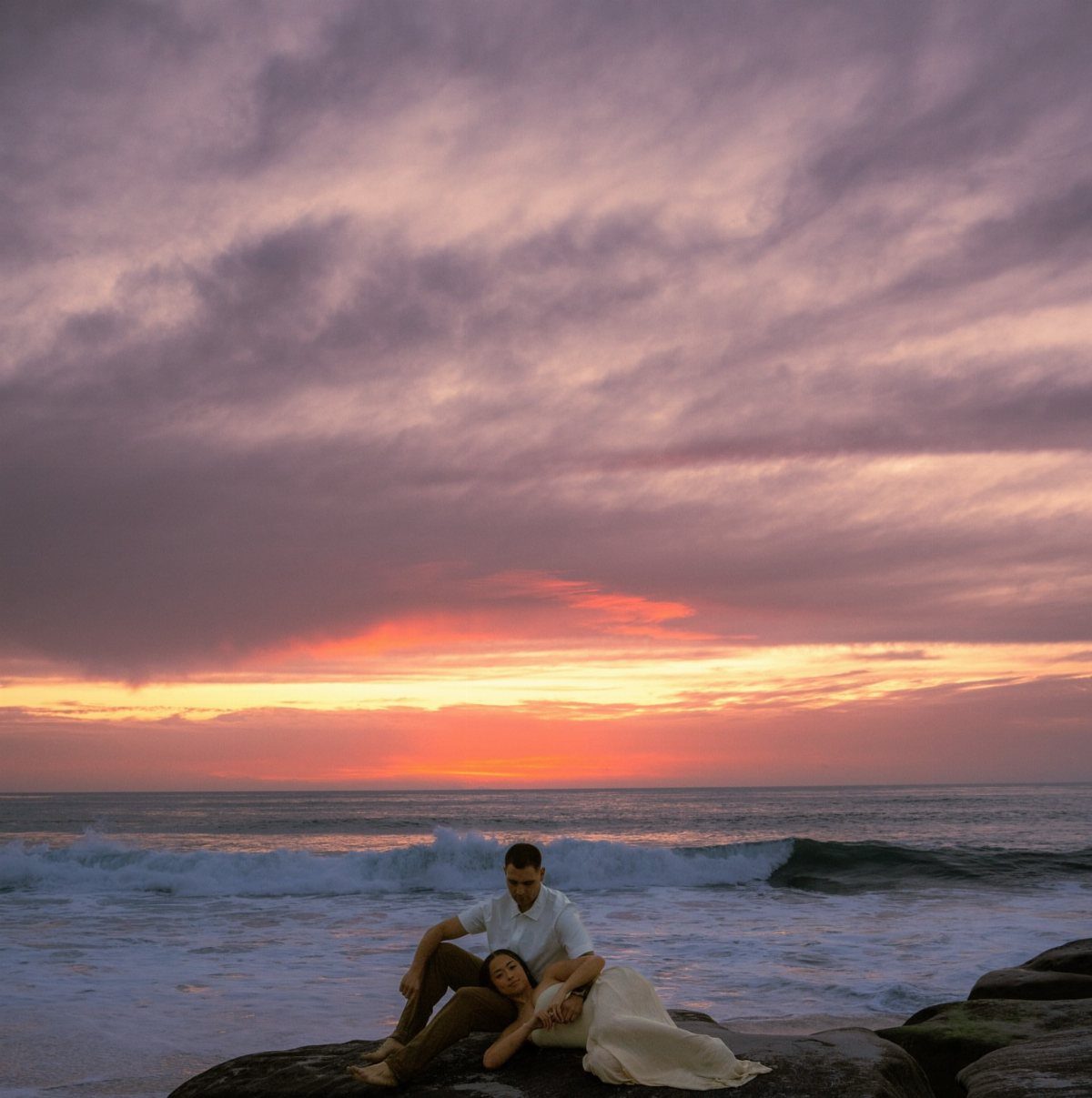 Couple sitting on coastal rocks during a vibrant purple and orange sunset beach scene