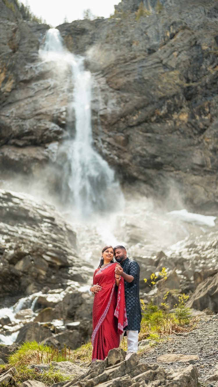 Couple in traditional Indian attire posing before a large waterfall and rocky cliffs.