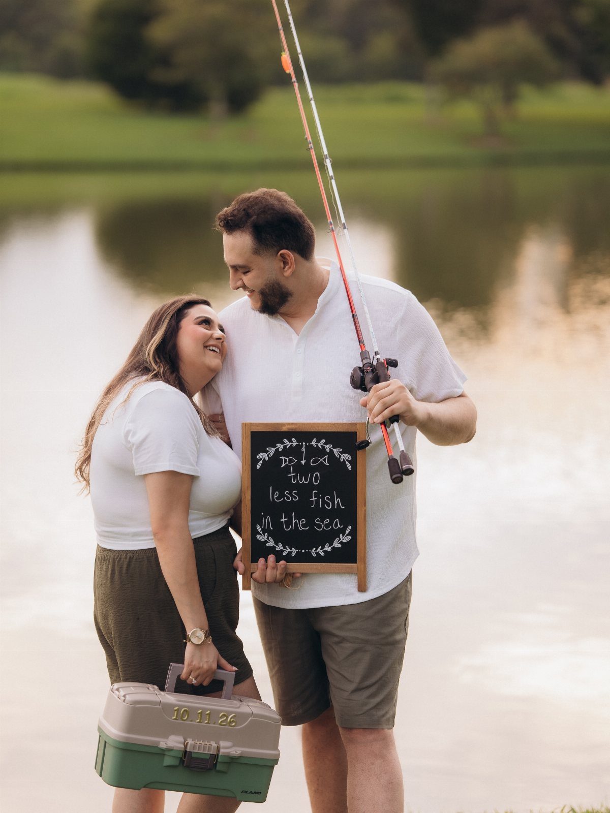Couple posing by a lake with a fishing rod and a chalkboard sign.