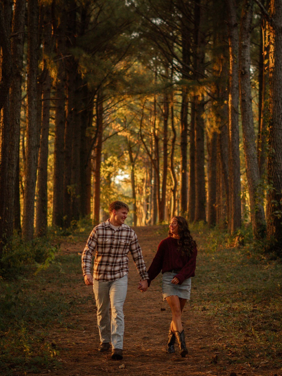 Couple walking hand-in-hand and smiling in a sunlit pine forest during golden hour.