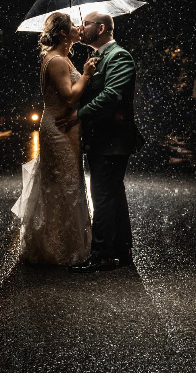 Couple in wedding attire kissing under an umbrella in the rain at night.
