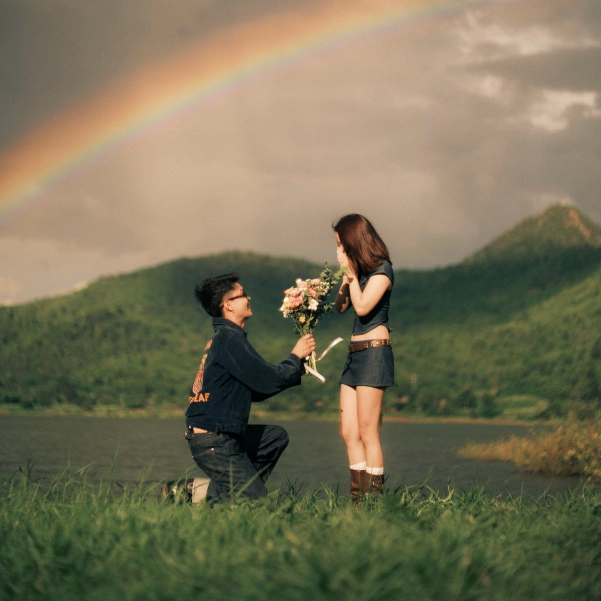 Man kneeling and giving flowers to a woman under a rainbow in nature.
