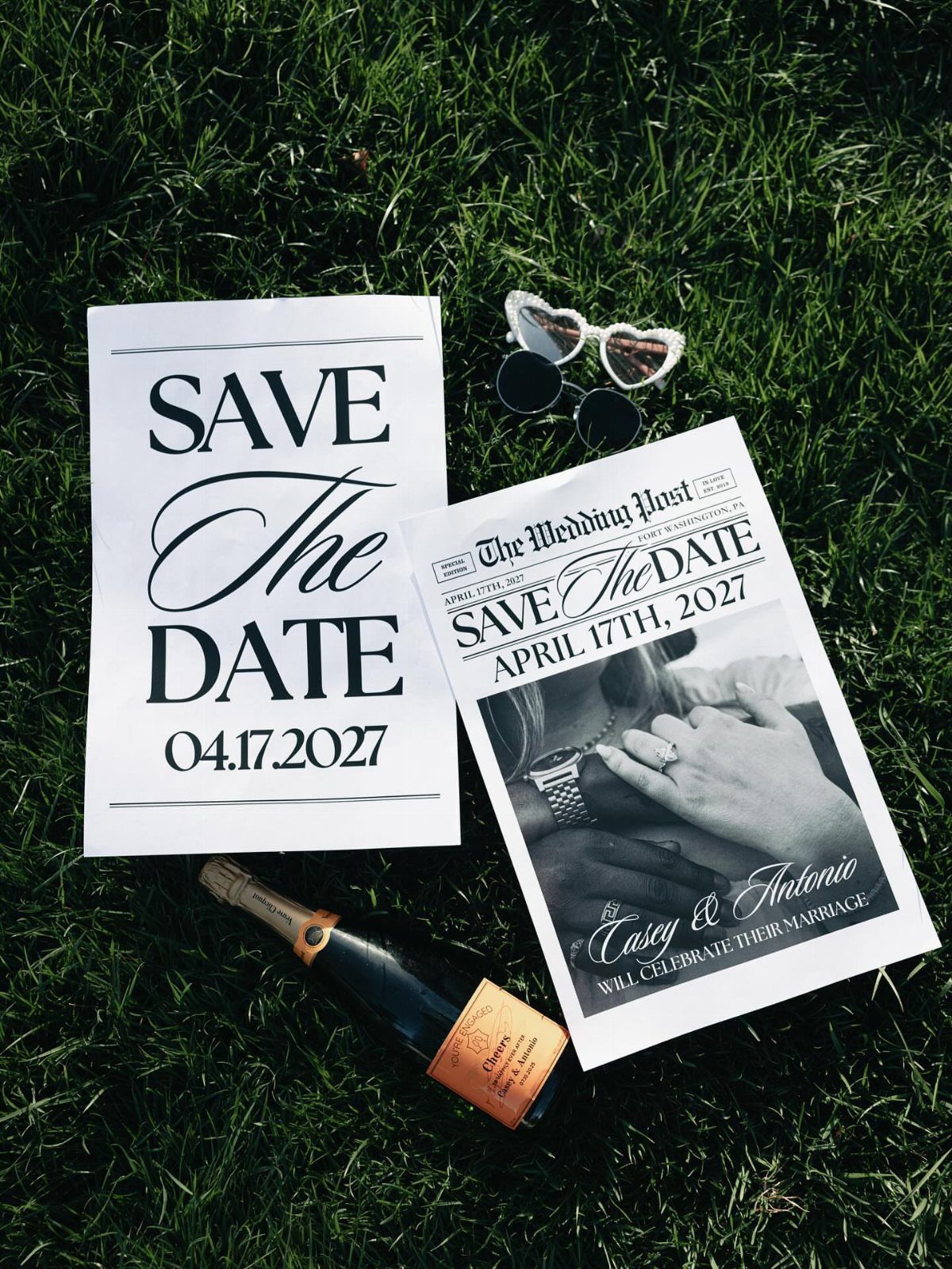 Save the date card, mock newspaper, champagne bottle, and sunglasses on green grass.