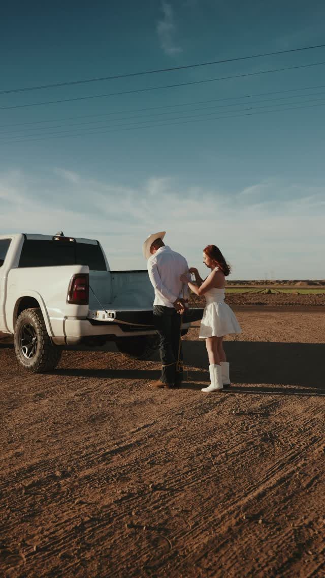 Couple in western attire standing by a white pickup truck on a dirt road.