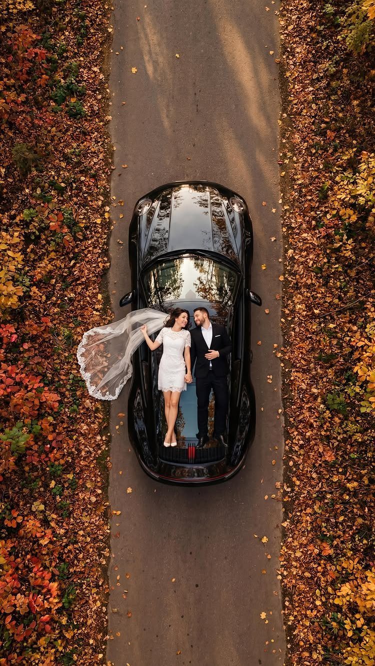 Couple in wedding attire standing on a black car amidst autumn leaves.