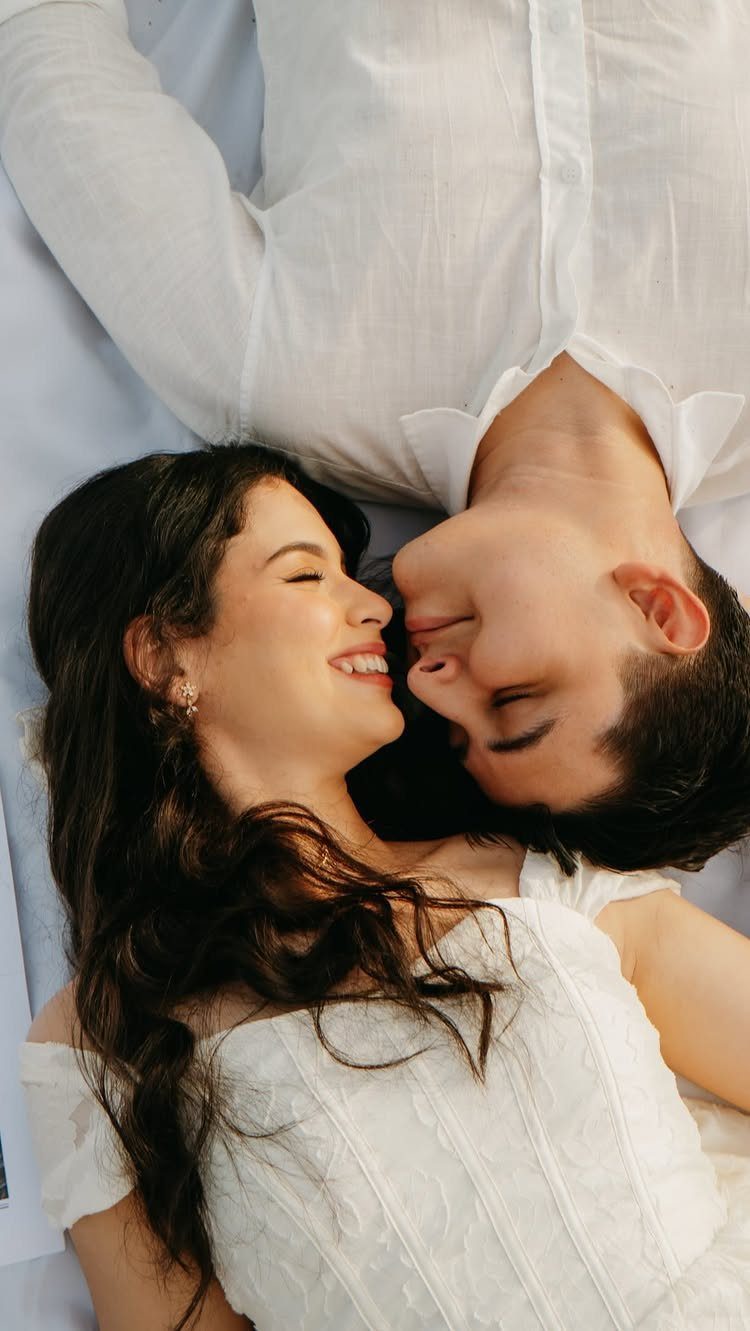 Couple in white clothing lying head-to-head and smiling at each other on white fabric.
