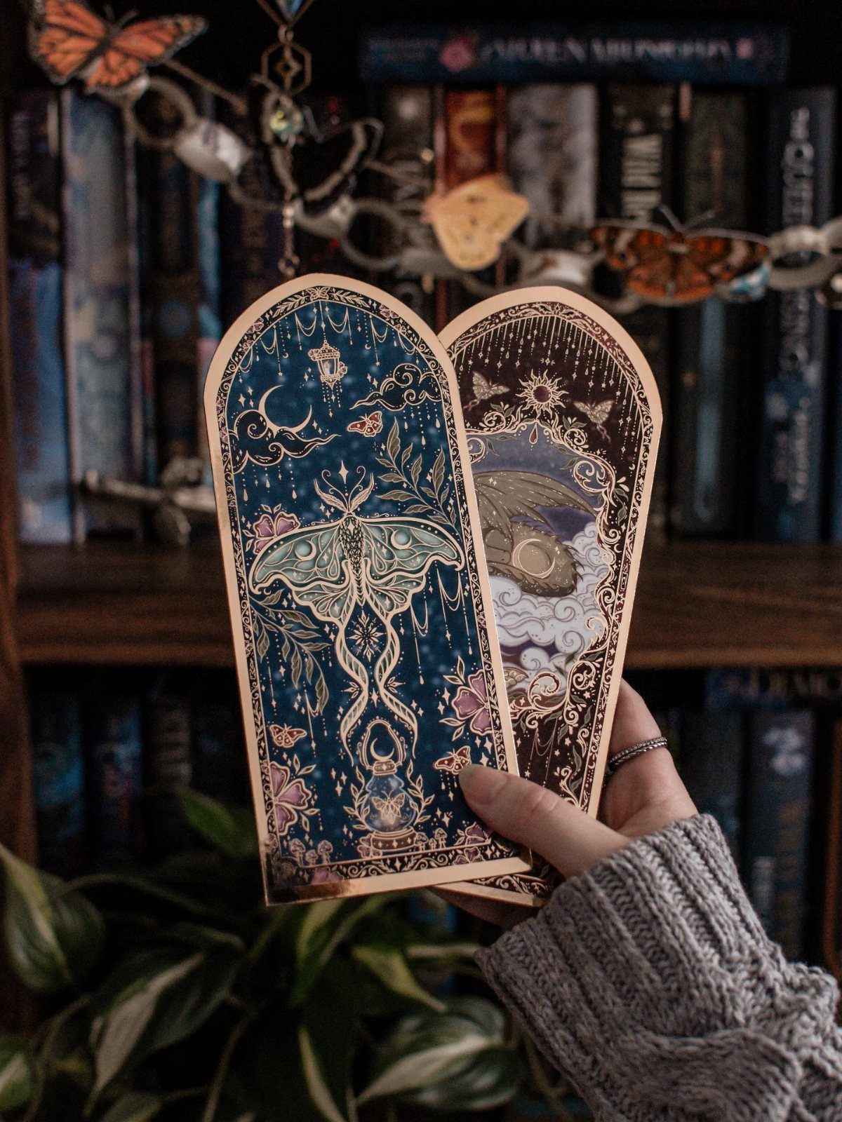 Ornate celestial bookmarks with moth illustrations held in front of a bookshelf.