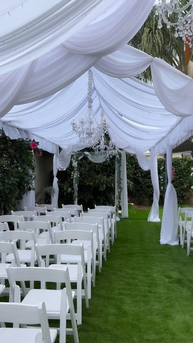 White folding chairs under a draped white canopy with chandeliers on a green lawn