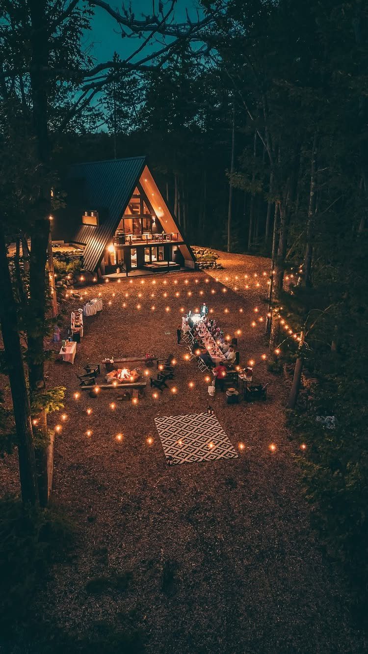 A-frame cabin with outdoor dining table and fire pit under string lights in woods