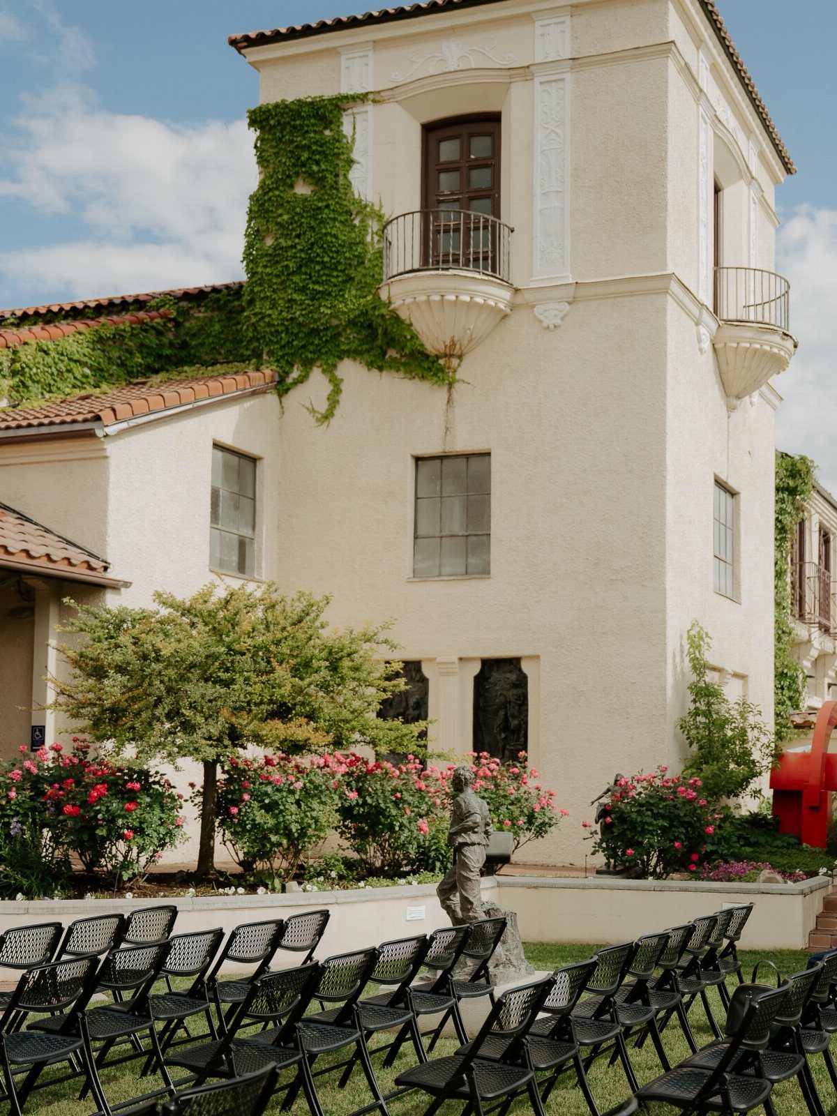 Black metal chairs arranged for a ceremony before a white Mediterranean building with ivy.