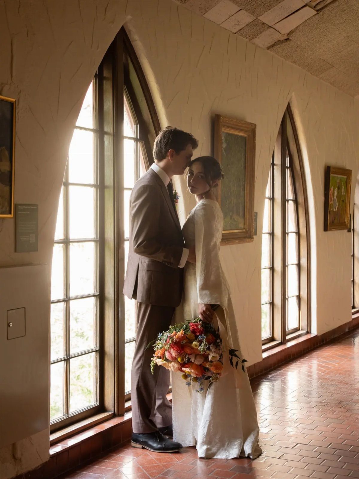 Bride and groom posing in an art gallery with arched windows and paintings.