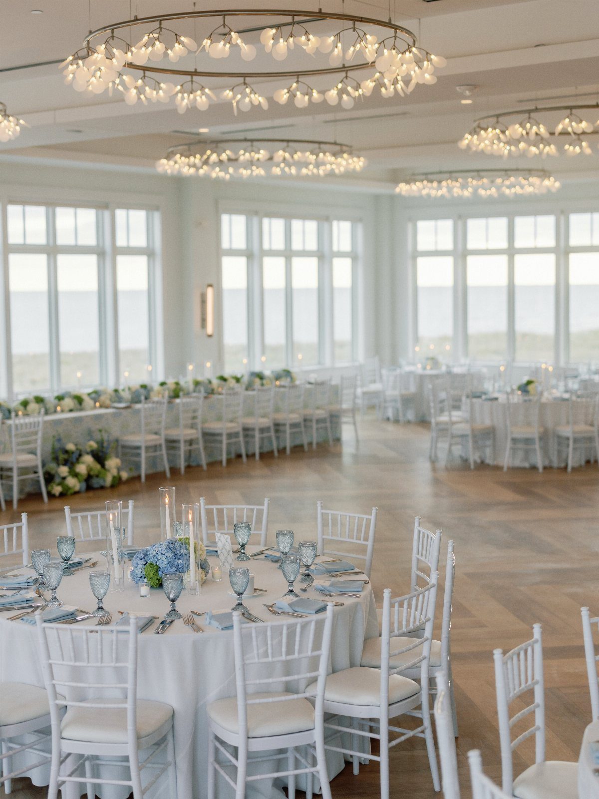 Wedding reception hall with white tables, blue accents, and beach views through windows.