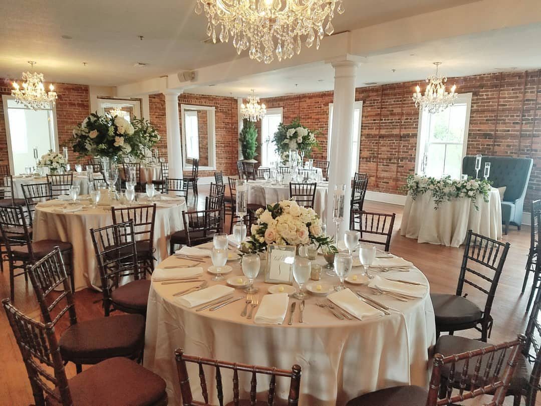 Wedding reception room with round tables, white flowers, and crystal chandeliers against brick walls.