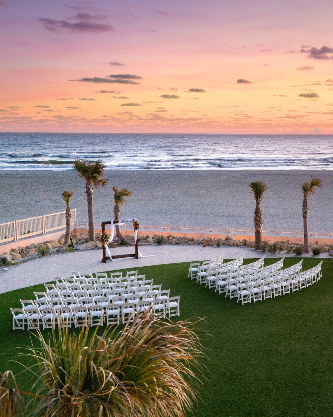 Wedding ceremony setup with white chairs and arch overlooking a beach at sunset.