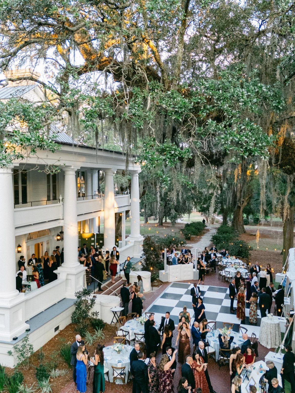 Outdoor wedding reception with guests on a checkered dance floor beside a white mansion.