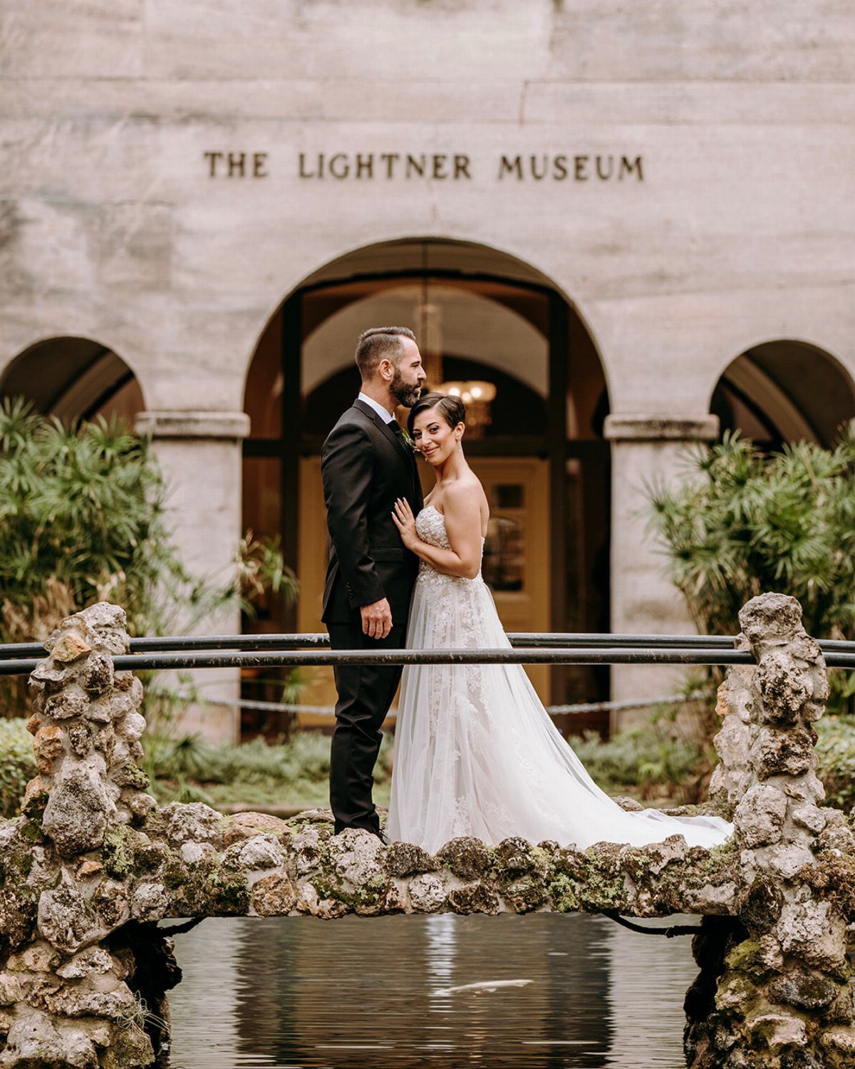Bride and groom posing on a stone bridge in front of The Lightner Museum.