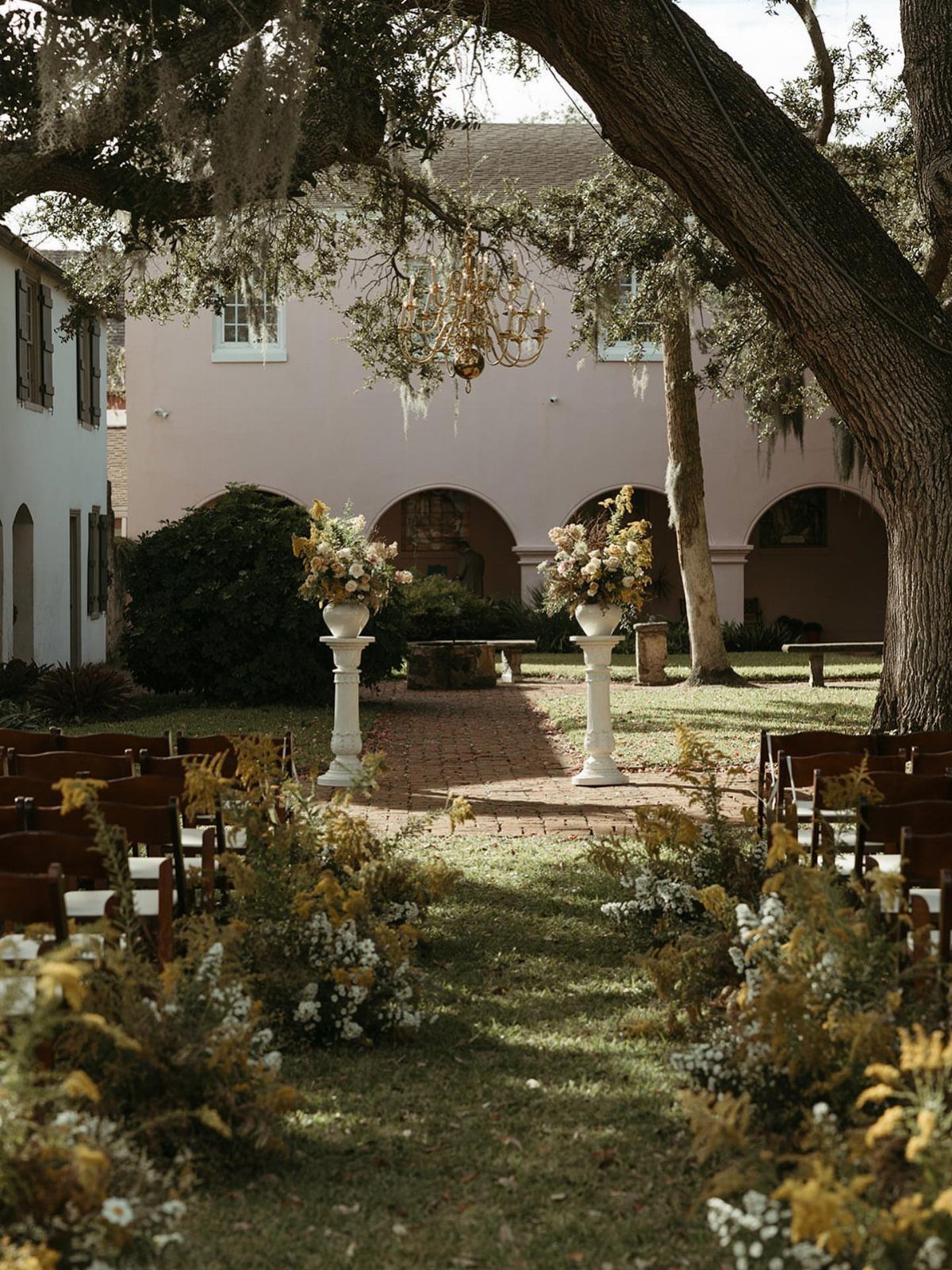 Outdoor wedding ceremony setup with floral pedestals and wooden chairs under an oak tree.