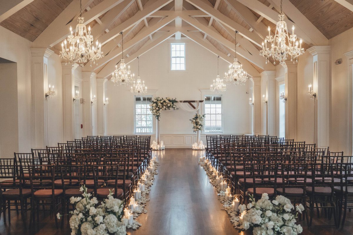 Wedding ceremony setup with white floral arrangements, candles, and crystal chandeliers in a hall.