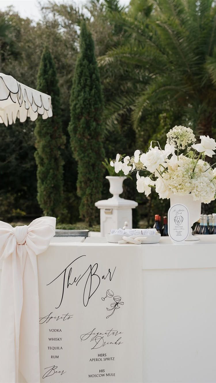 White bar sign with cursive lettering and signature drinks next to white flowers.