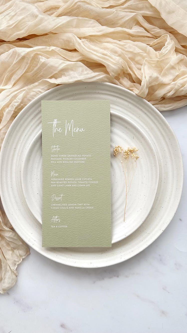 Sage green menu card on a white plate with dried flowers and beige fabric.