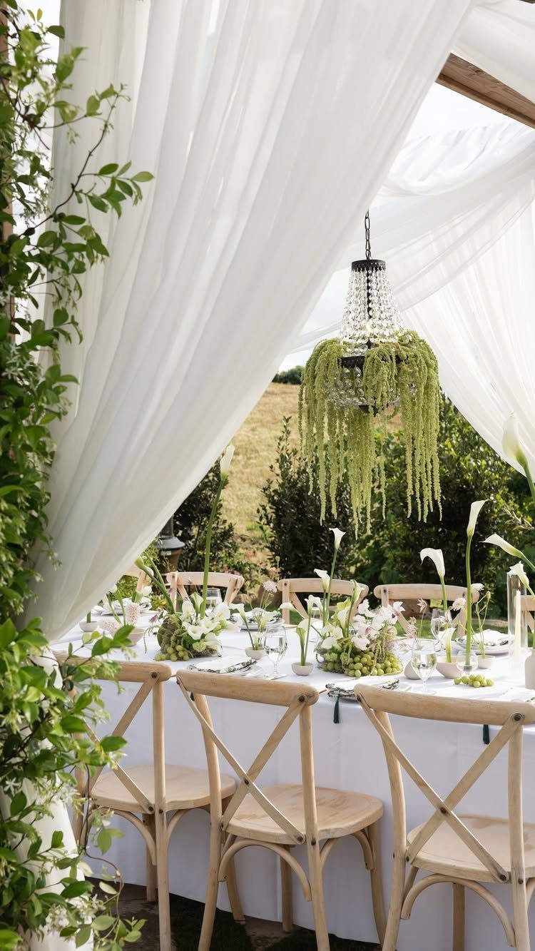 Outdoor wedding reception table with white linens, cross-back chairs, and draped white fabric canopy.