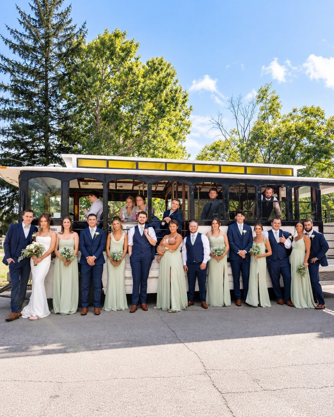 Wedding party posing in front of a white trolley bus outdoors with trees.