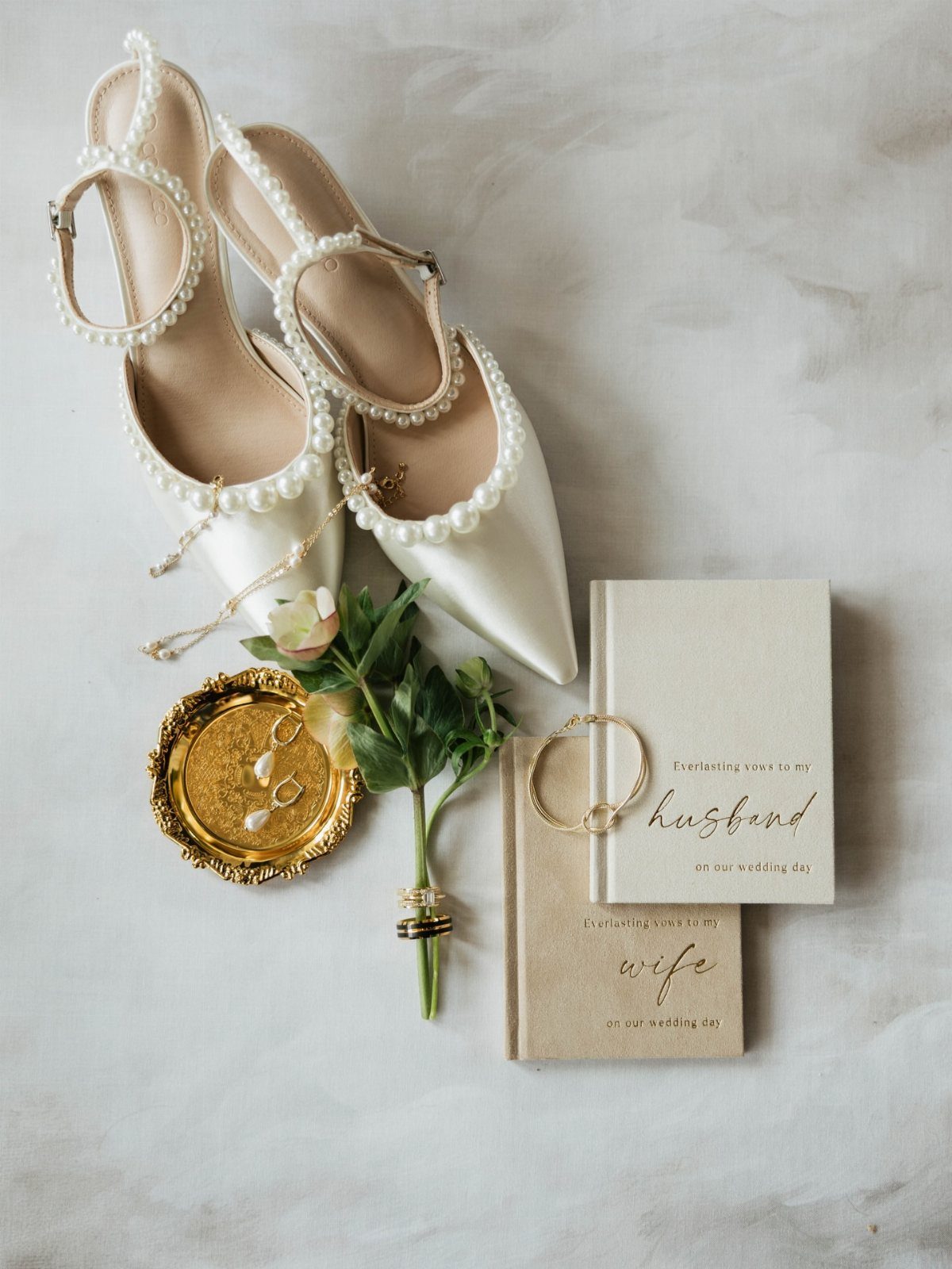White pearl bridal shoes, wedding notebooks, and gold jewelry on a grey background.