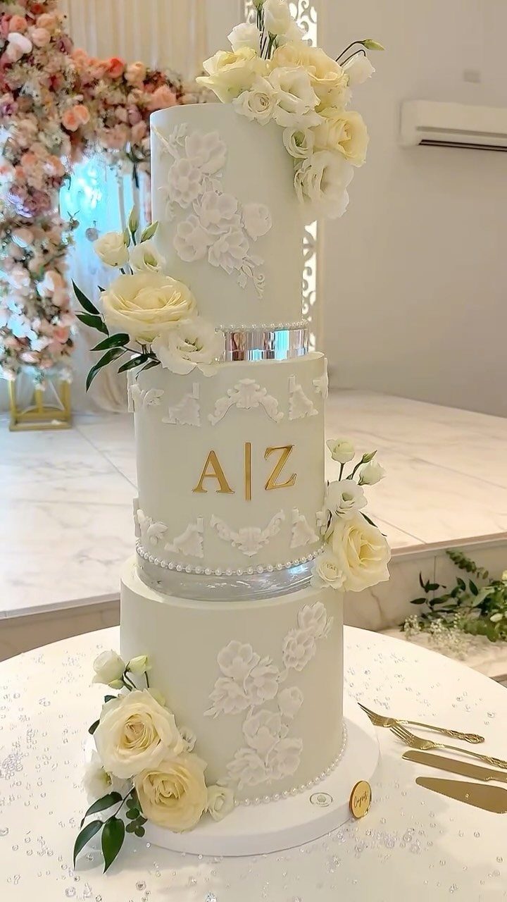 Three-tier white wedding cake with cream roses and gold initials on a table.