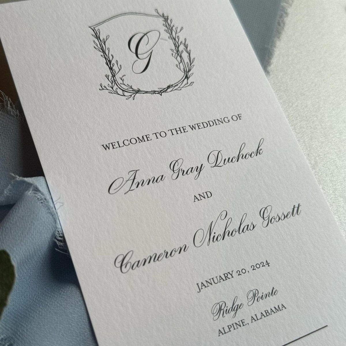 Wedding invitation card with elegant black calligraphy on white paper and blue fabric.