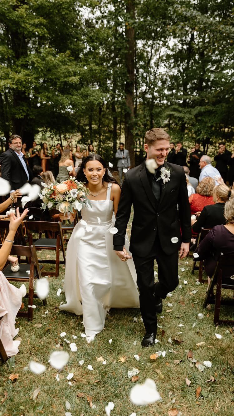 Bride and groom walking down a grassy aisle with white petals falling around them.