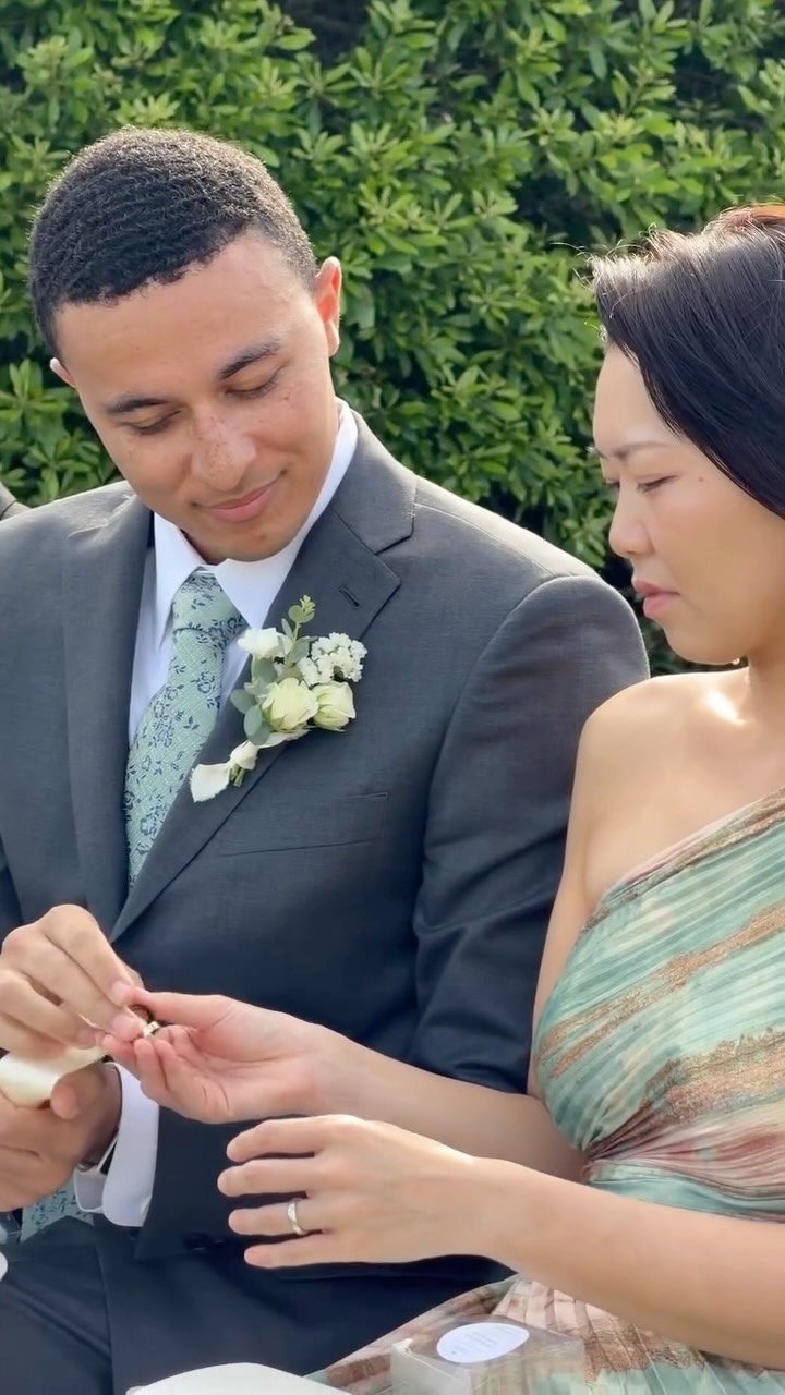 Groom in grey suit placing a wedding ring on the bride's finger outdoors.