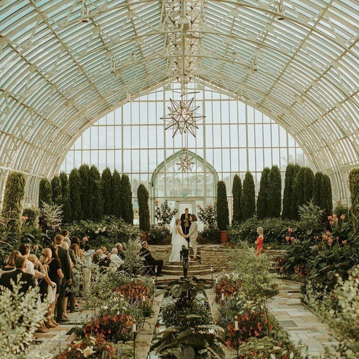 Wedding ceremony inside a glass conservatory filled with lush gardens and green hedges.