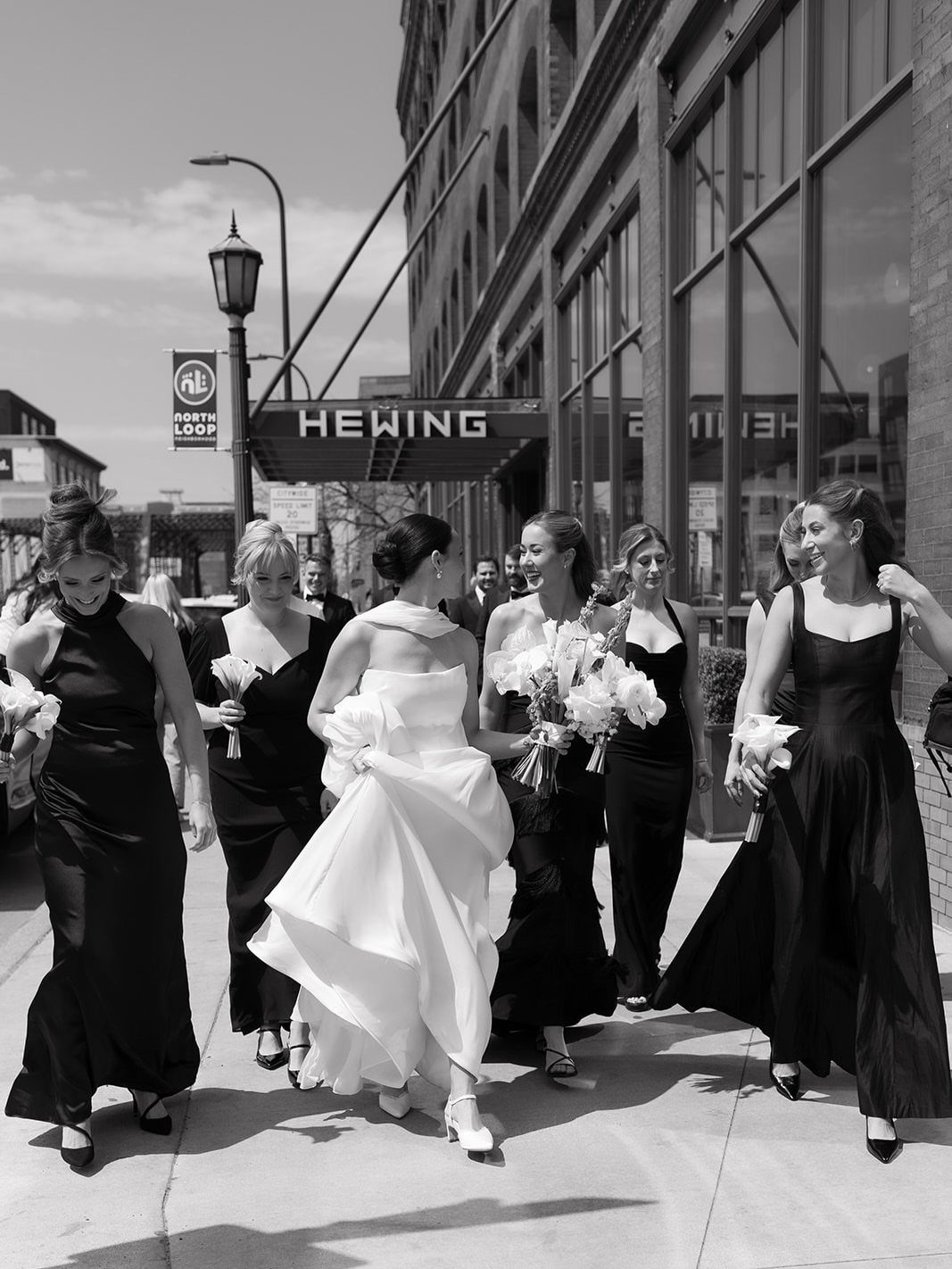 Bride and bridesmaids walking on a sidewalk in front of the Hewing Hotel.