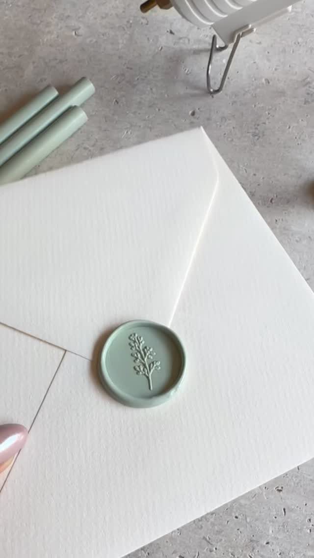 White textured envelope with a sage green botanical wax seal on grey concrete.