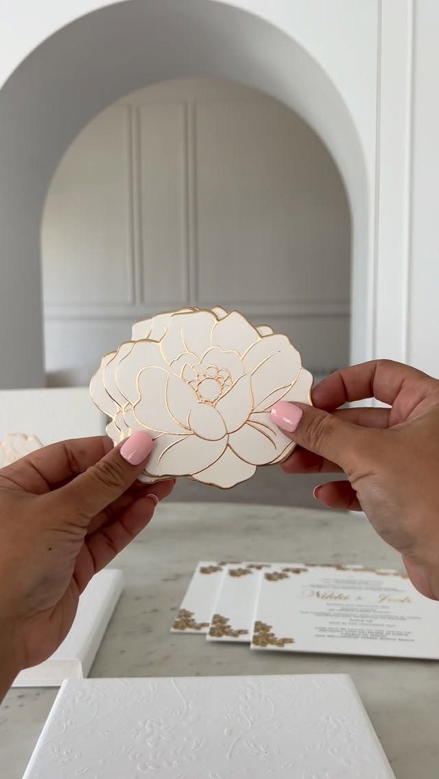 Flower-shaped white card with gold foil outlines held over gold foil wedding invitations.