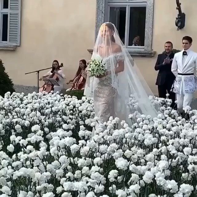 Bride in white gown and veil walking alone through white flowers at wedding