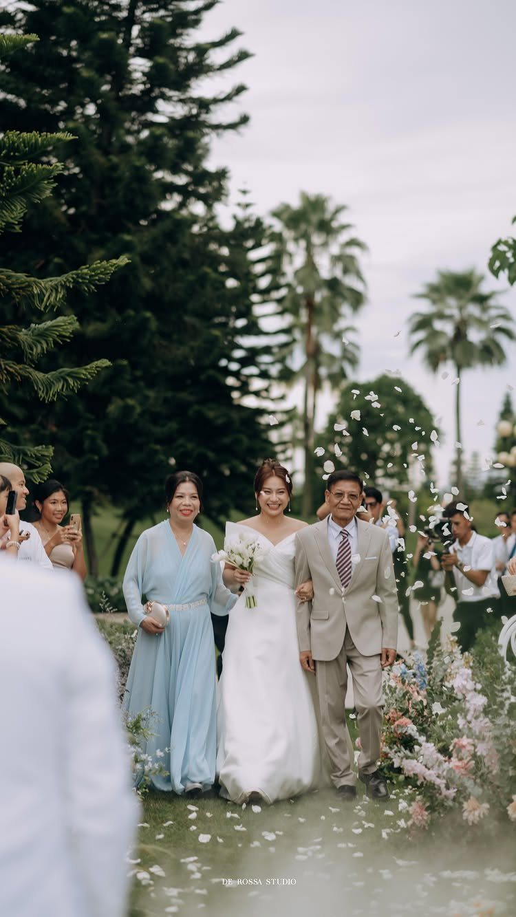 Bride in white gown walking down the aisle escorted by both parents outdoors.