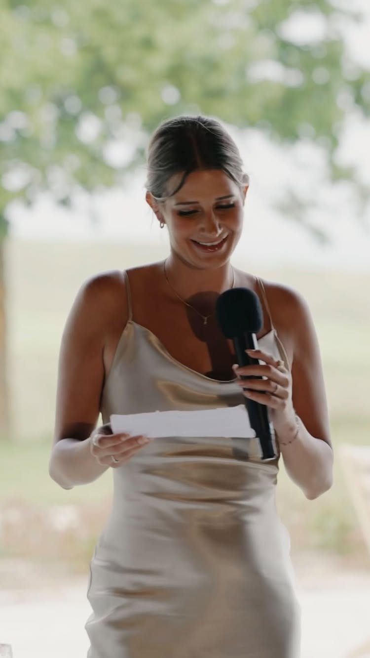 Woman in champagne satin dress speaking into a microphone while reading a note.