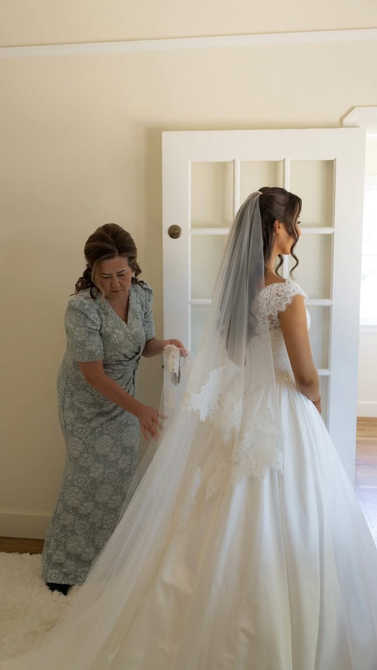 Woman helping a bride put on her white wedding dress and veil indoors.