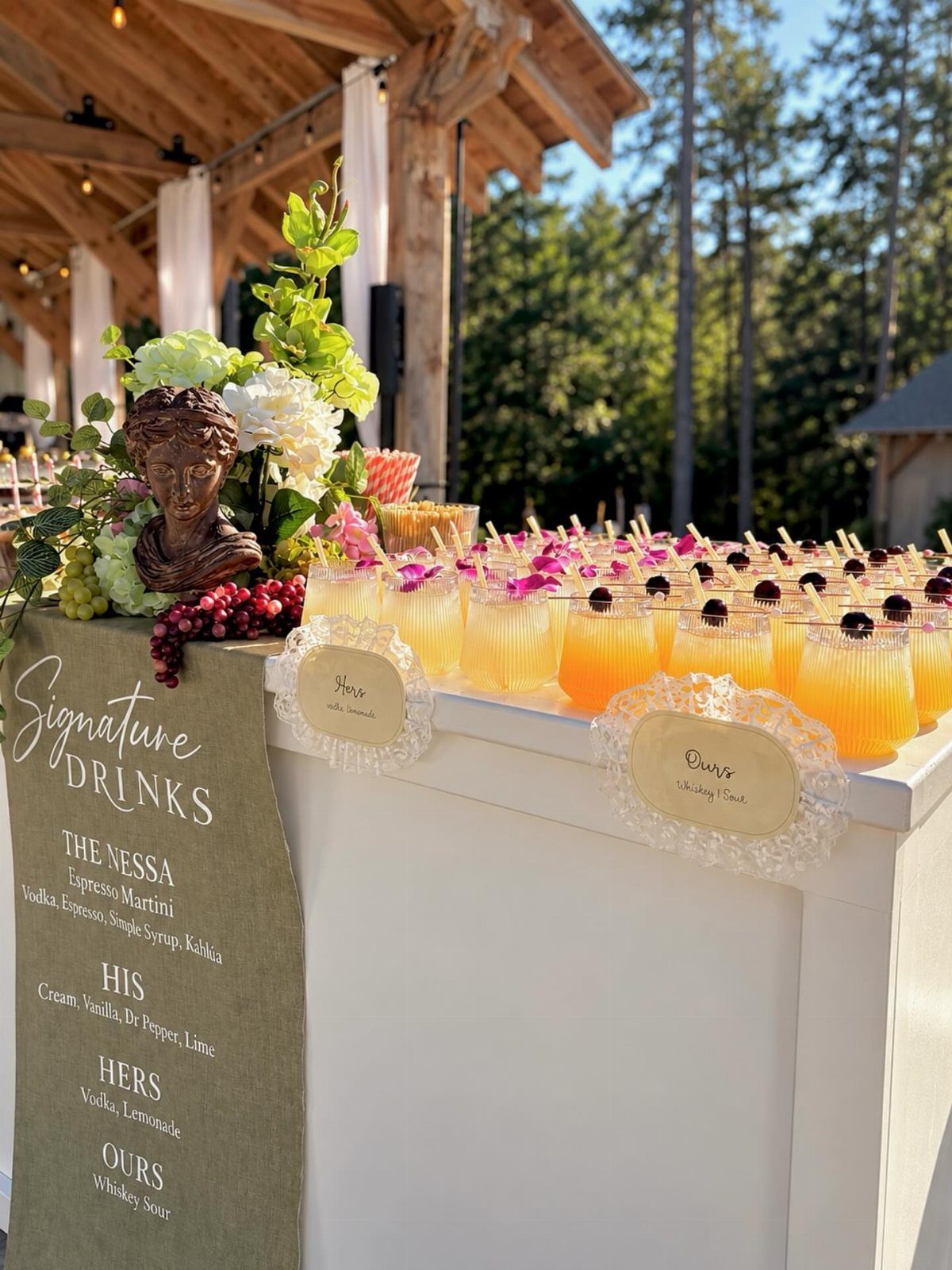 Yellow signature cocktails with berries on a white bar at an outdoor wedding.