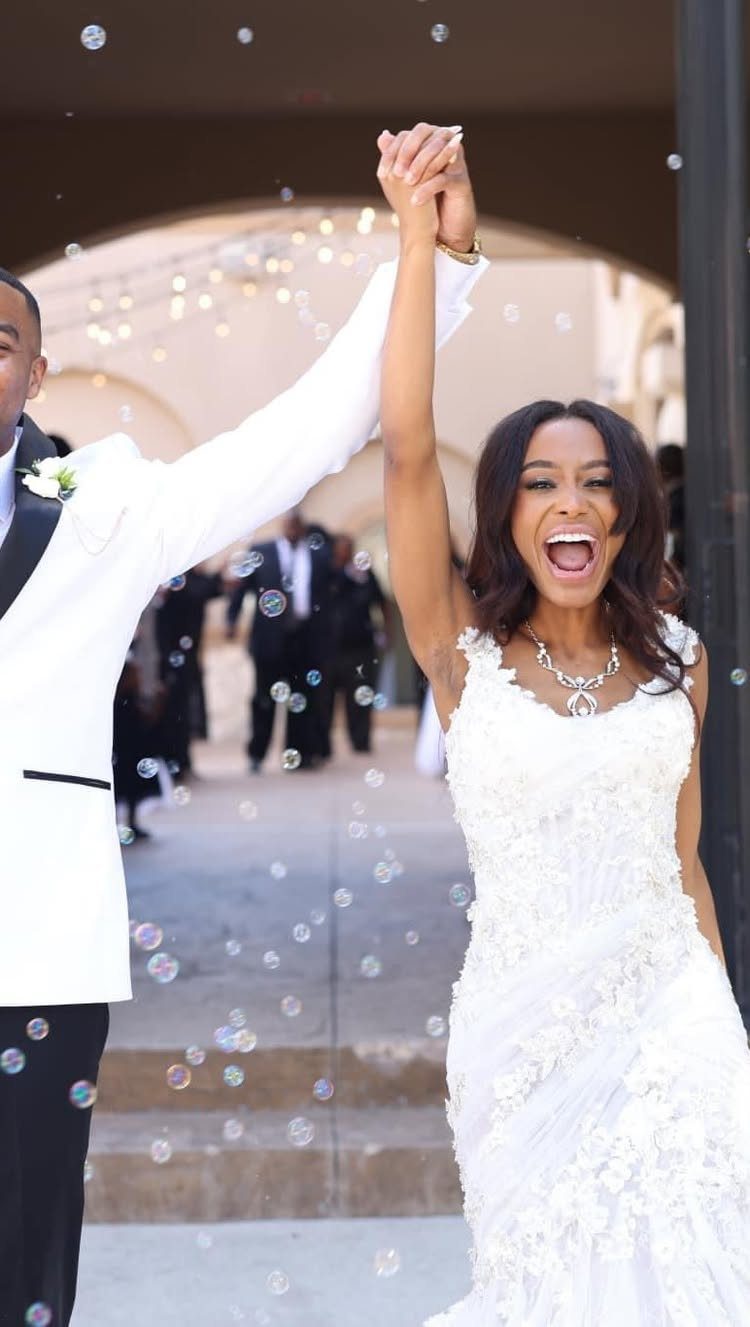 Bride and groom cheering with raised hands while walking through bubbles at wedding.